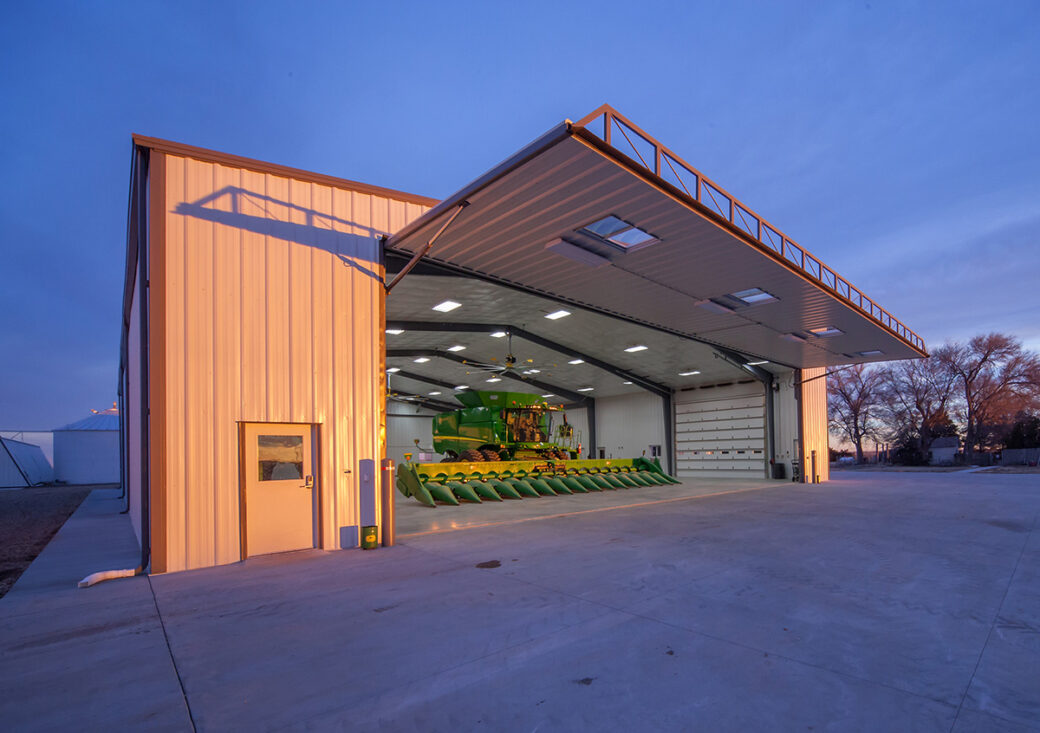 Agricultural metal building, hydraulic door open with large green combine inside building, twilight